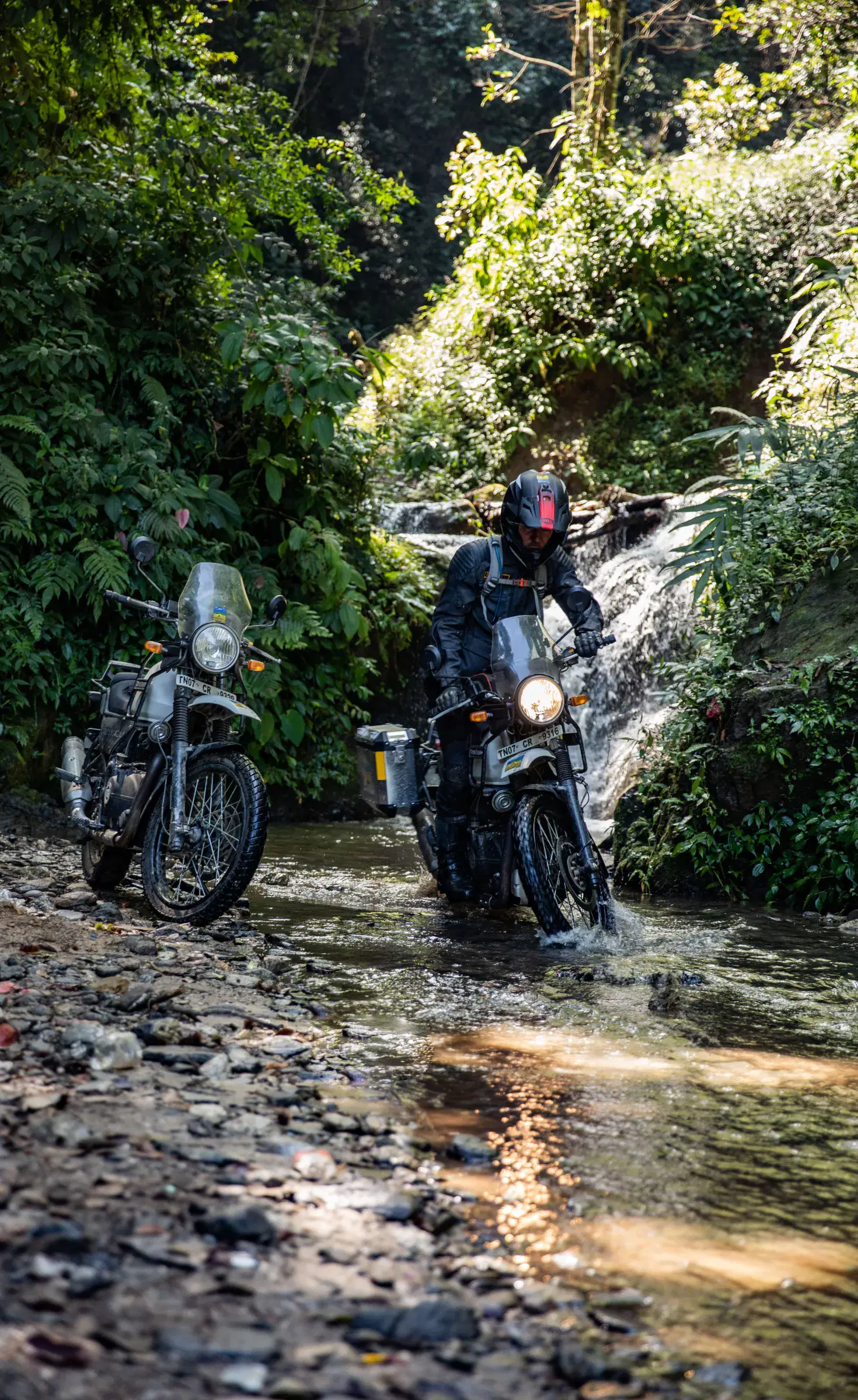 Portrait-oriented Outdoor adventure scene featuring people, a vehicle, rocky terrain, a built structure, and greenery.
