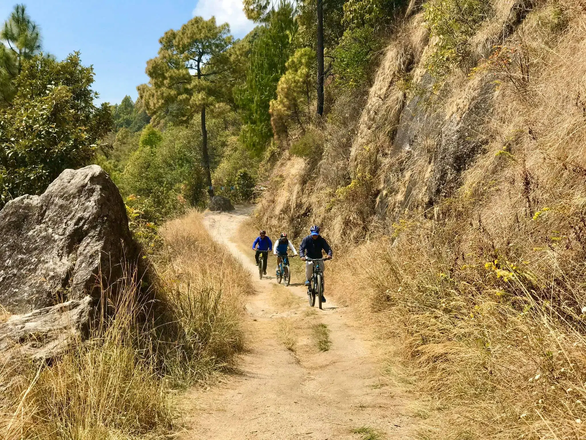 Outdoor scene featuring people, rocky terrain, a built structure, blue sky, and greenery.