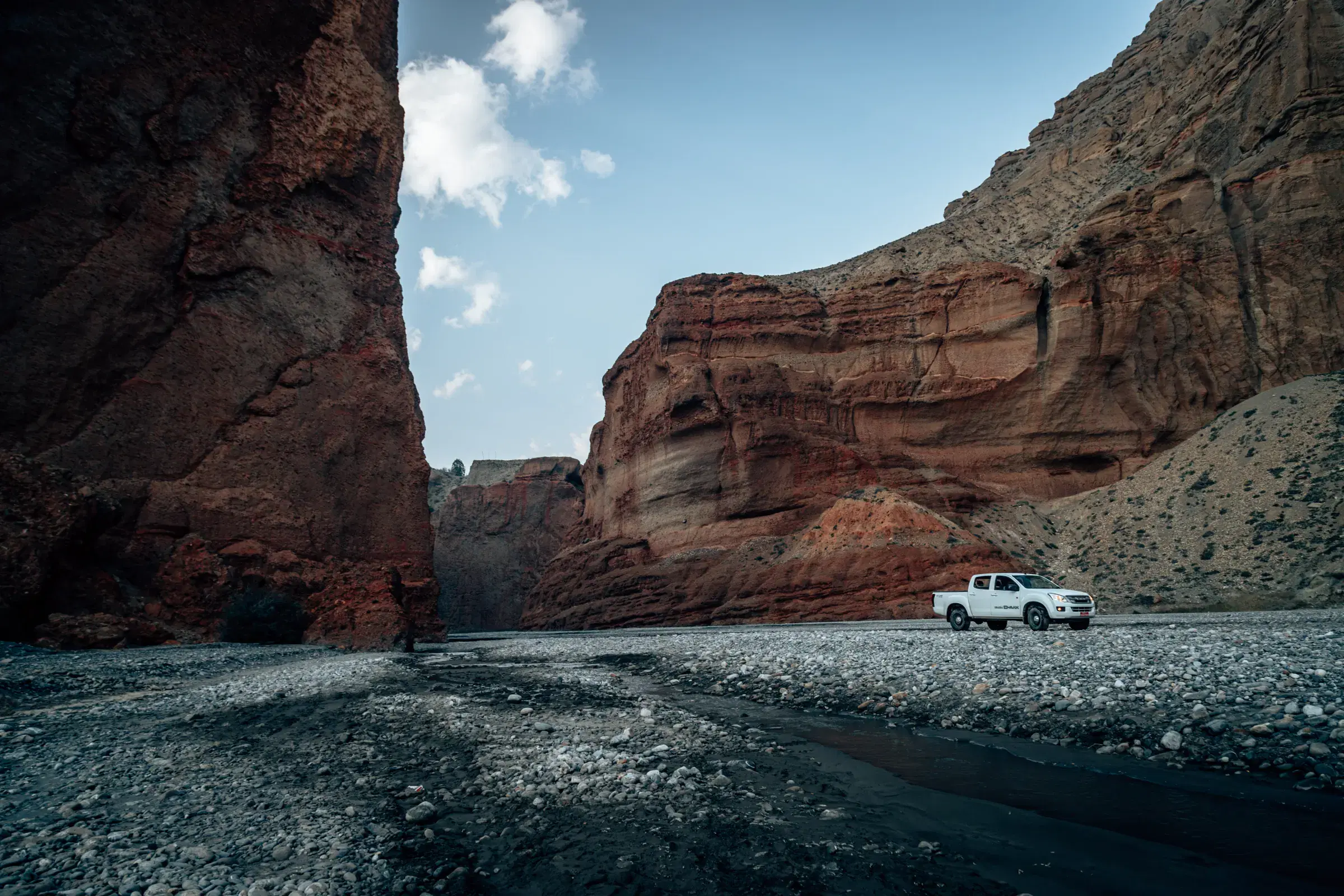 Outdoor vehicle scene featuring a vehicle, rocky terrain, a built structure, and cloudy sky.